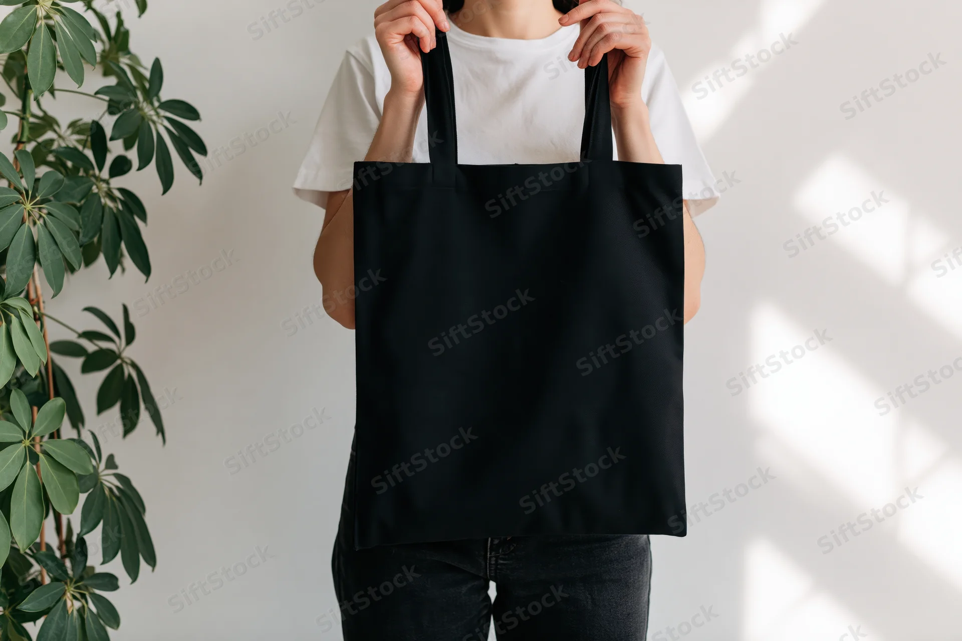 Woman holding plain black canvas tote bag in front of white wall with plant and sunlight shadows