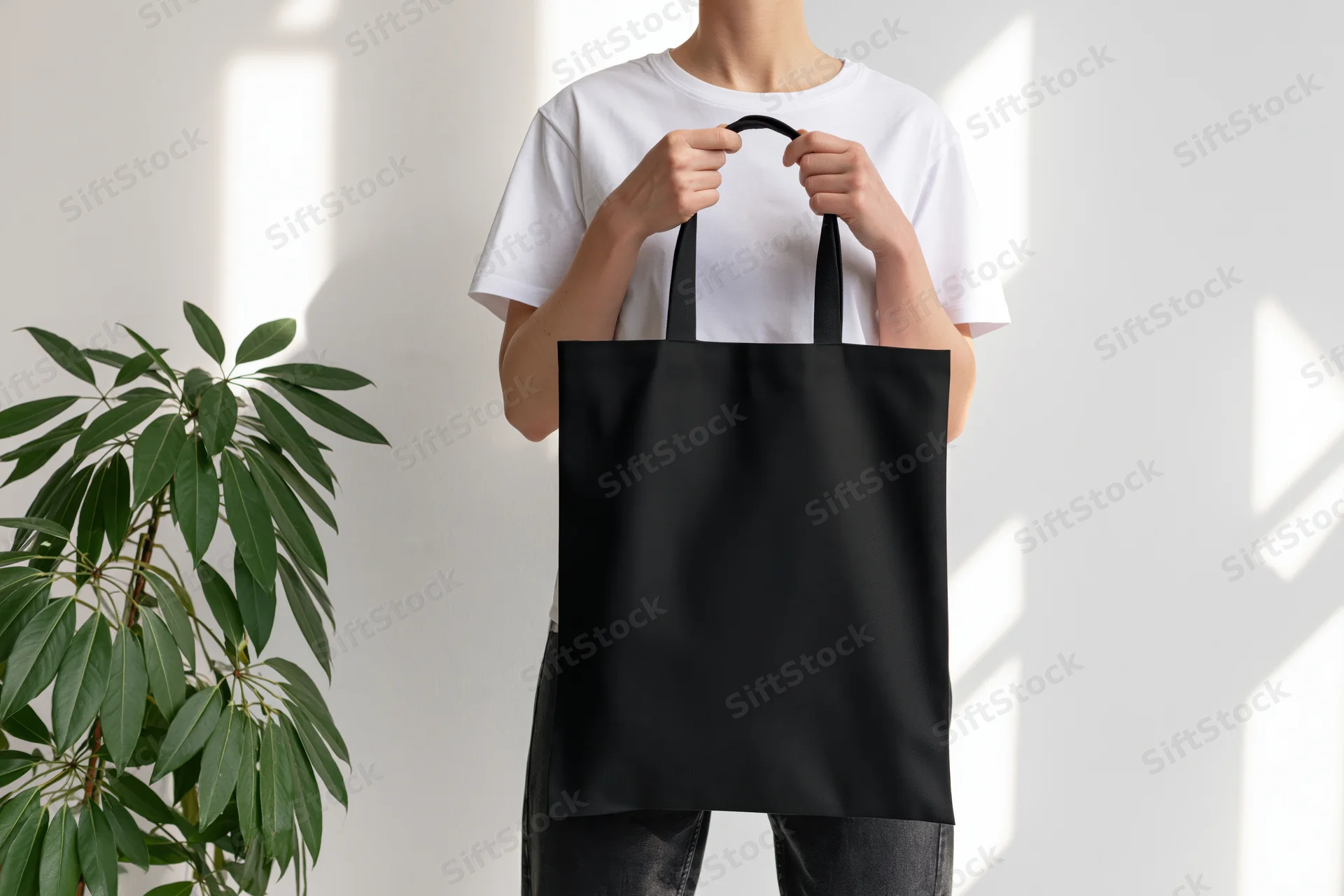 Woman holding plain black canvas tote bag in front of white wall with plant beside her