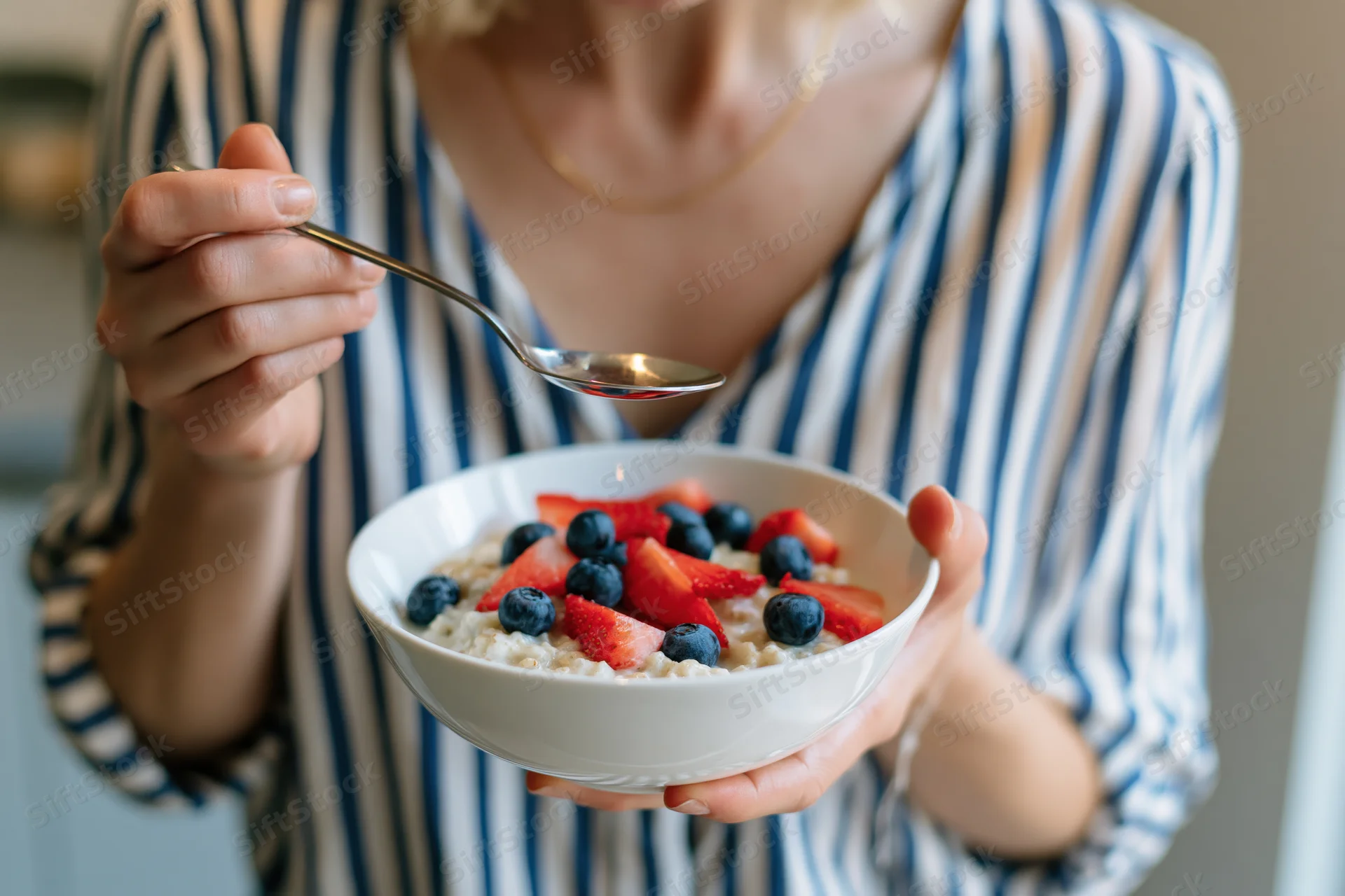 Woman holding a bowl of oatmeal topped with strawberries and blueberries while eating with a spoon in a kitchen setting