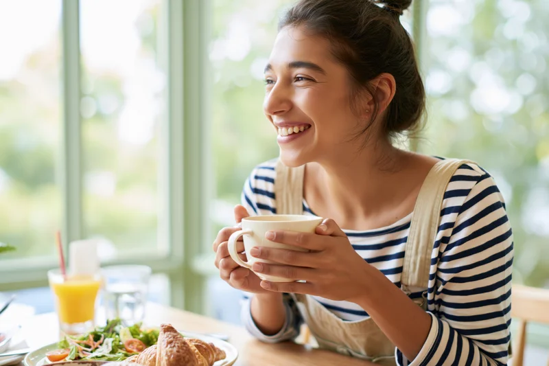 Young woman smiling while holding a white ceramic mug at a breakfast table by a window