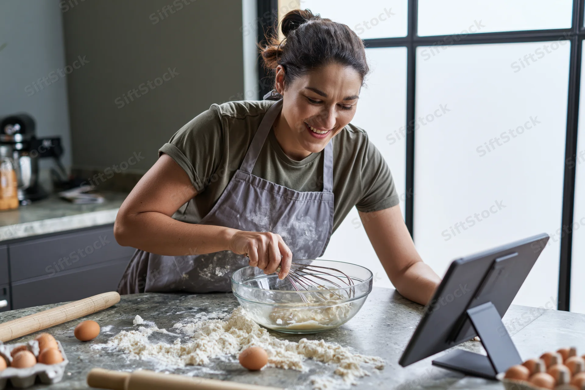 Woman whisking dough in a glass bowl in a modern kitchen while following a recipe on a tablet