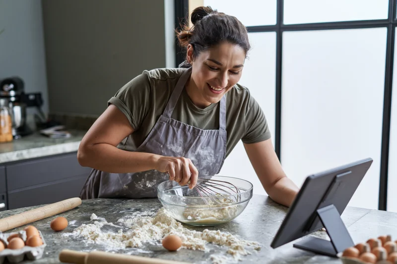 Woman whisking dough in a glass bowl in a modern kitchen while following a recipe on a tablet