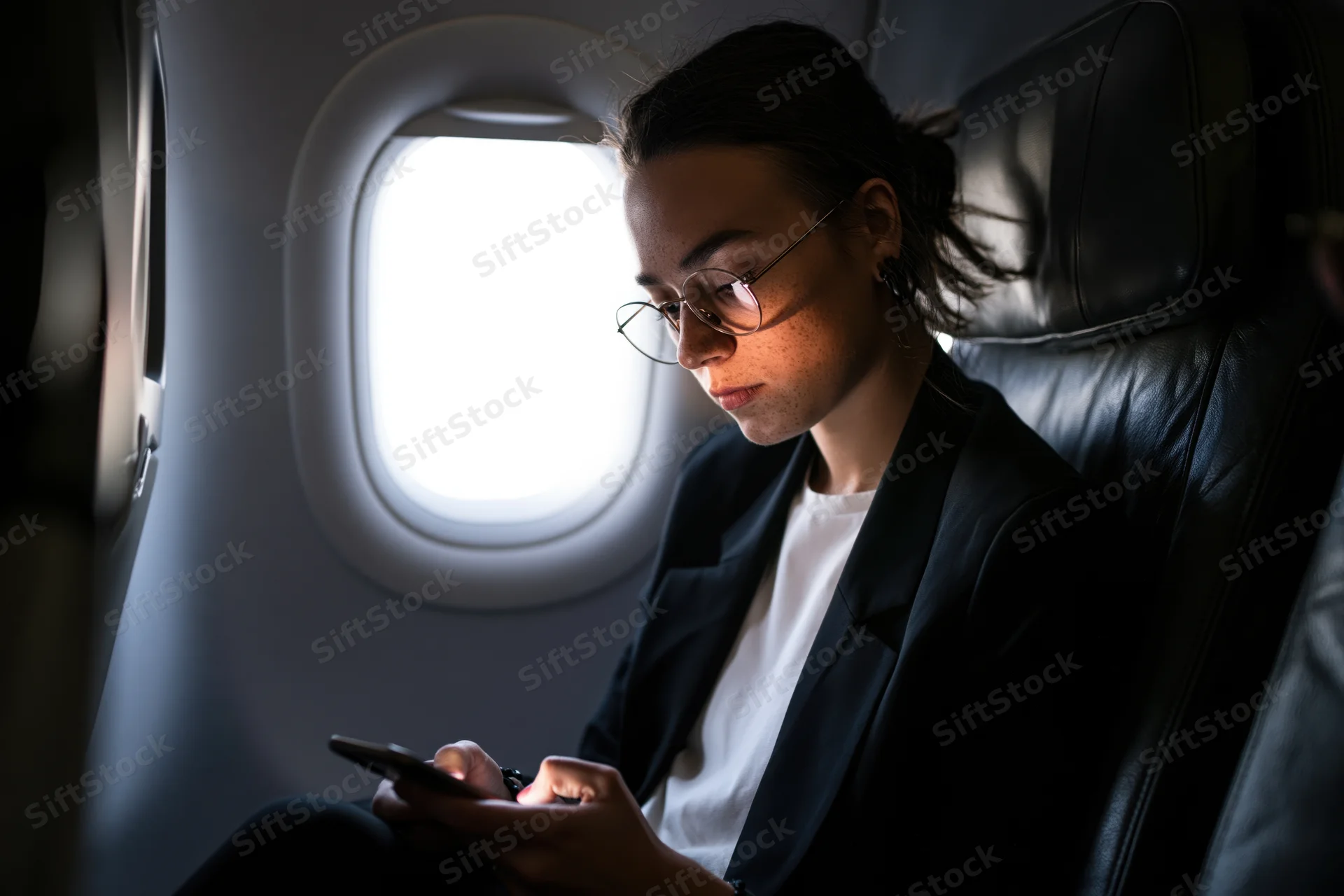 Woman in round glasses focused on smartphone while seated by airplane window