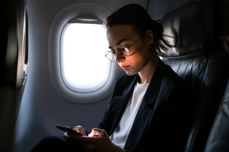 Woman in round glasses focused on smartphone while seated by airplane window