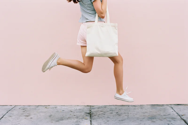 Woman mid-jump holding cream canvas tote bag on pink wall sidewalk