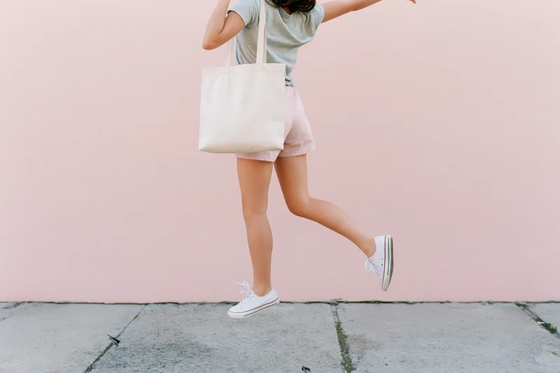 Woman mid-jump holding a cream canvas tote bag on a pink wall sidewalk