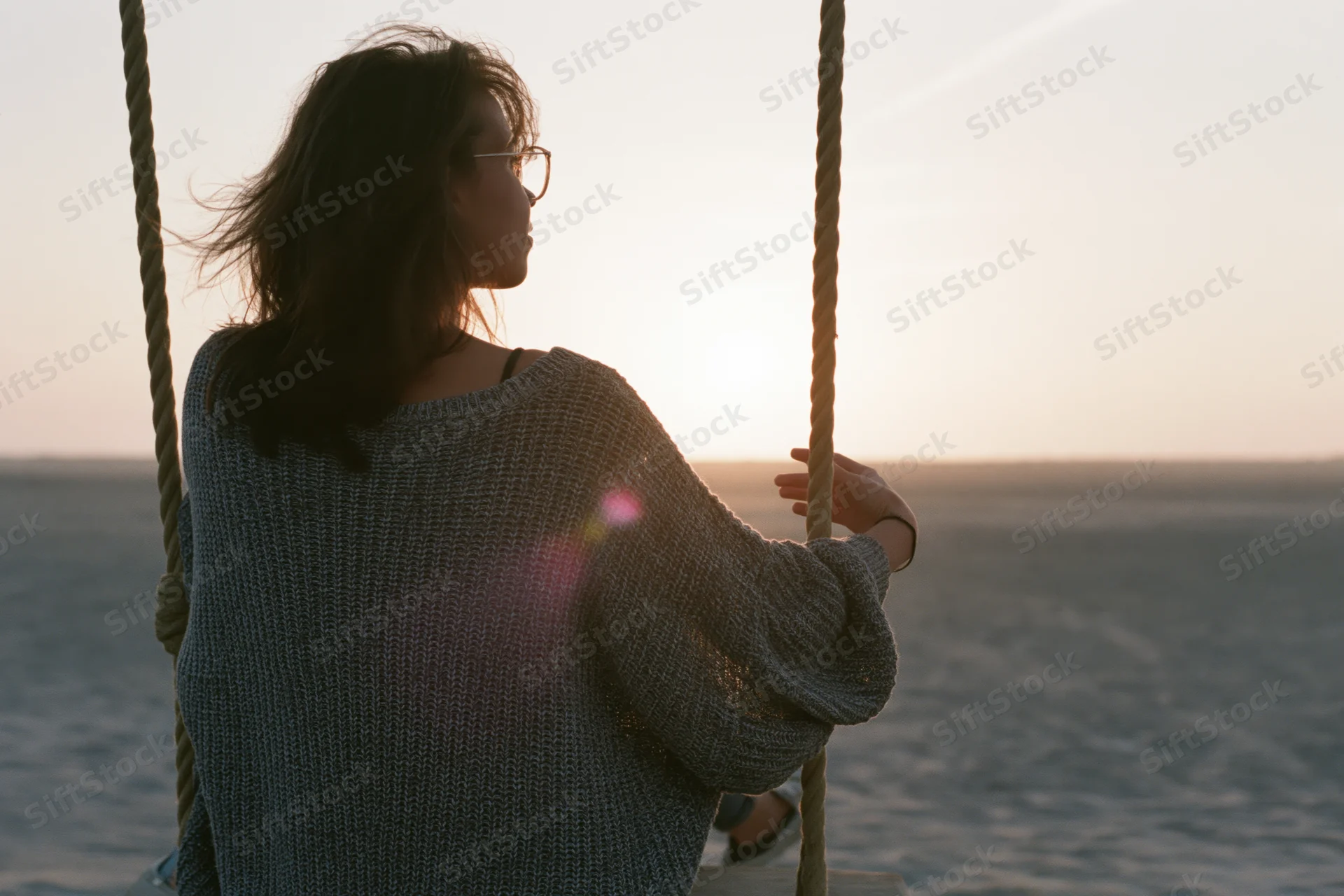 Young woman sitting on a rope swing facing the ocean at sunset