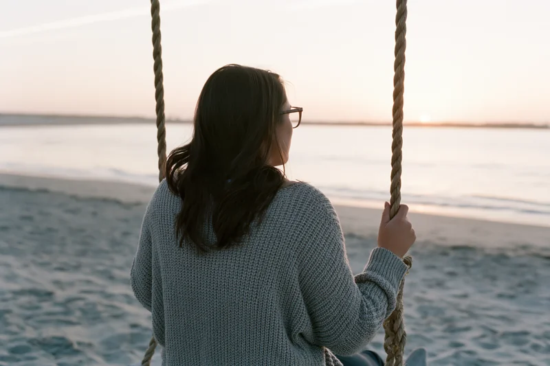 Woman sitting on rope swing facing ocean at sunset on sandy beach