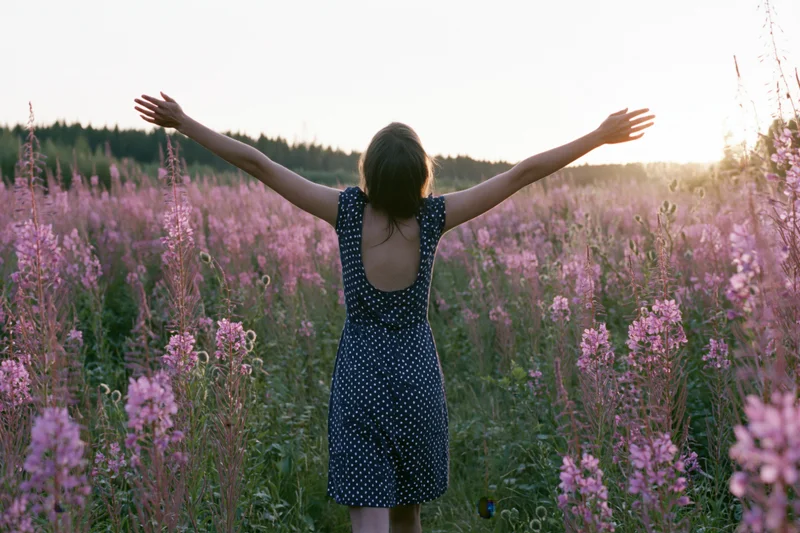 Woman with arms raised standing in a field of pink fireweed flowers at sunset