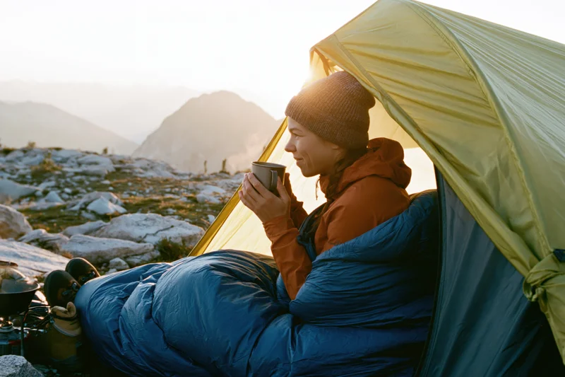 Woman in blue sleeping bag sipping coffee from mug at tent entrance in mountain sunrise