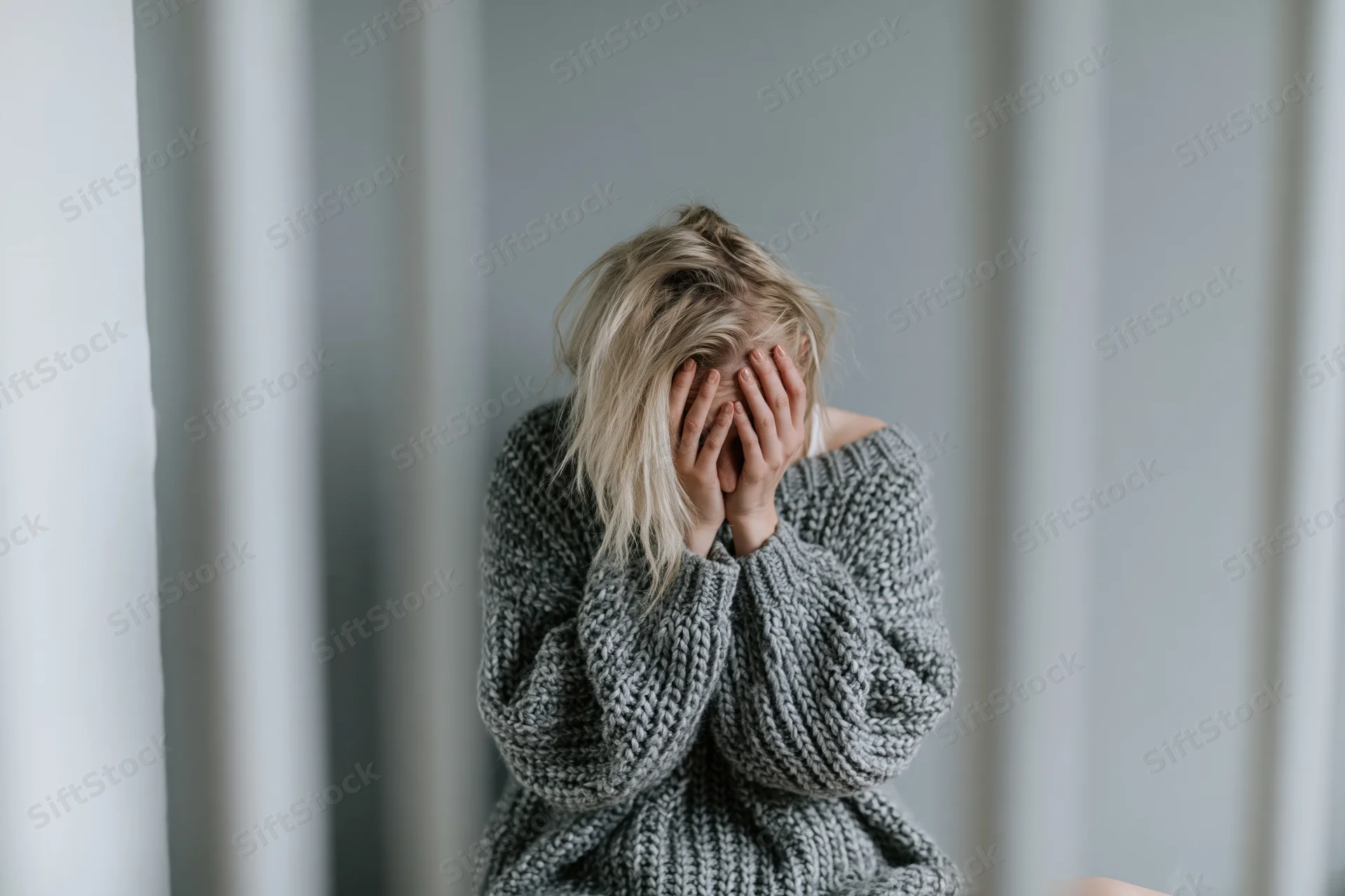 Young woman sitting on floor against gray wall covering face with hands in distress