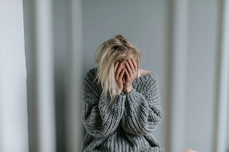 Young woman sitting on floor against gray wall covering face with hands in distress