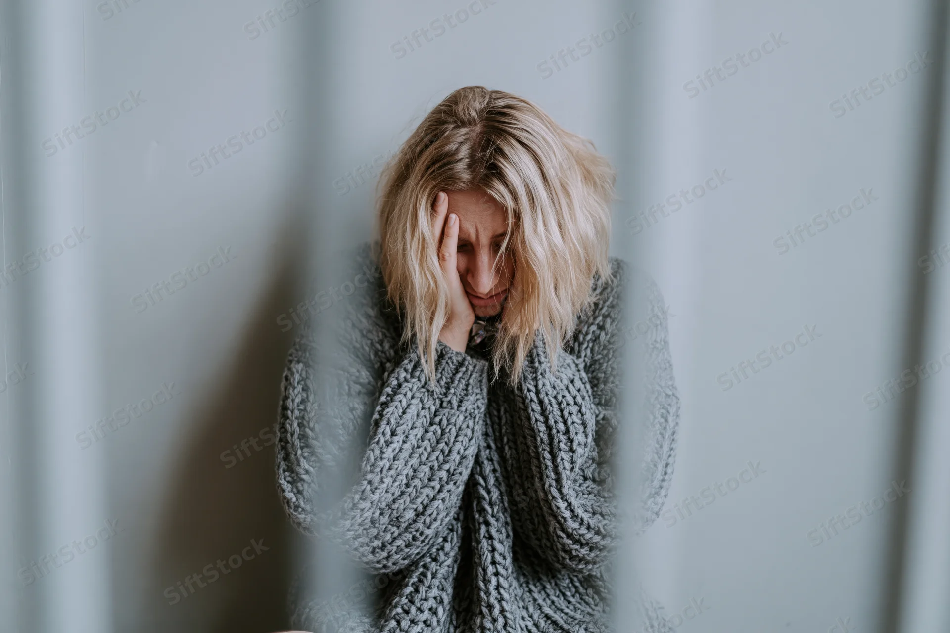 Young woman sitting on floor against grey wall with hand on face, wearing grey knitted sweater