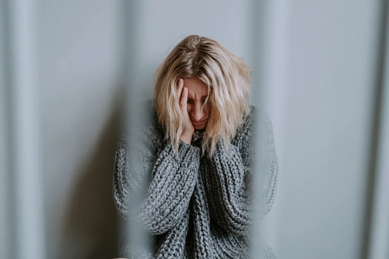 Young woman sitting on floor against grey wall with hand on face, wearing grey knitted sweater