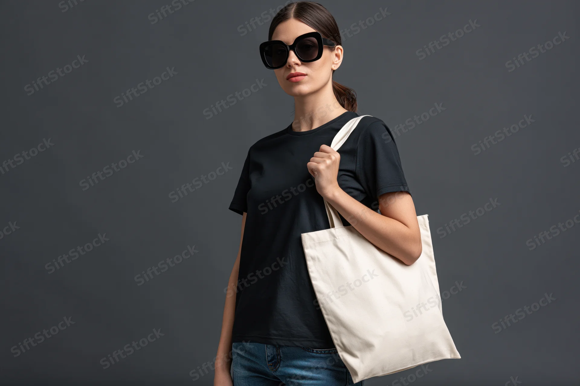 Woman in black tshirt and jeans holding white tote bag against dark grey studio background