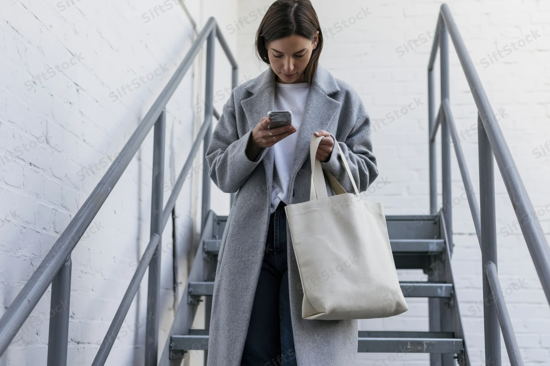 Woman walking down metal staircase while checking smartphone, carrying tote bag