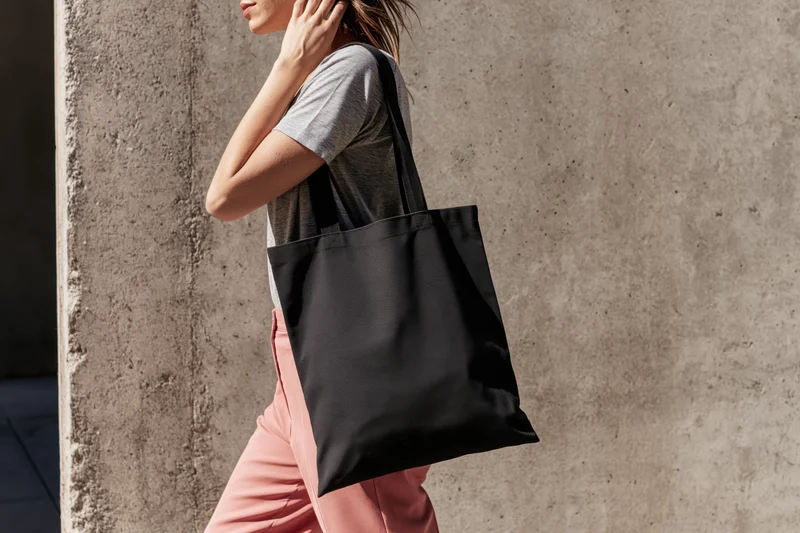 Woman walking outdoors with black tote bag over shoulder near concrete wall
