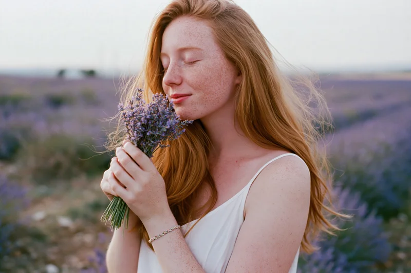 Young woman with long auburn hair smelling a small bouquet of lavender in a field