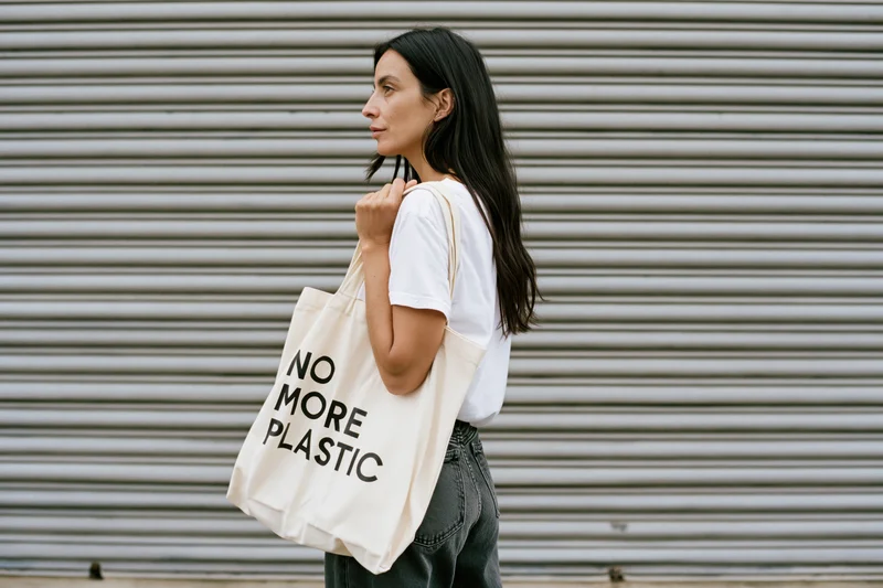 Young woman in profile holding a tote bag with no more plastic text against a metal shutter