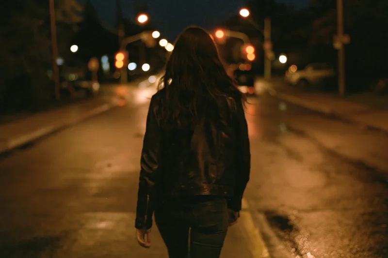 Woman walking away on wet city street at night, backview in leather jacket