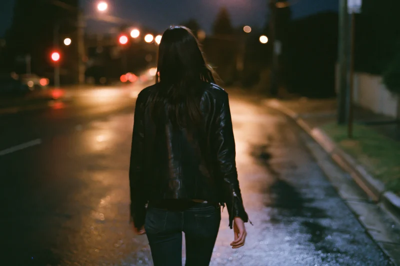 Woman walking away on wet city street at night, wearing black leather jacket and jeans, under streetlights and traffic signals