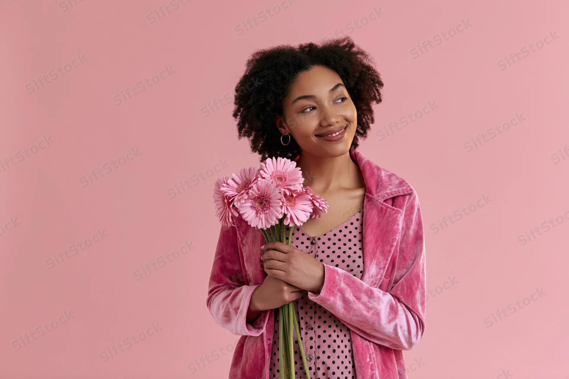 Young black woman holding pink gerbera flowers against pink background, smiling and looking away