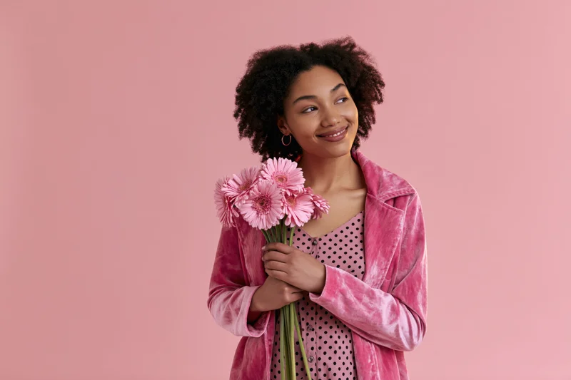 Young black woman holding pink gerbera flowers against pink background, smiling and looking away