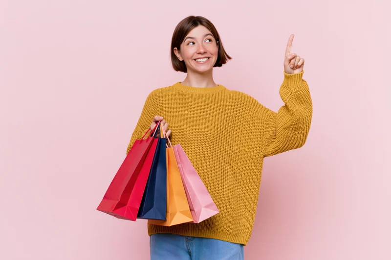 Woman holding colorful shopping bags and pointing upward against pink background