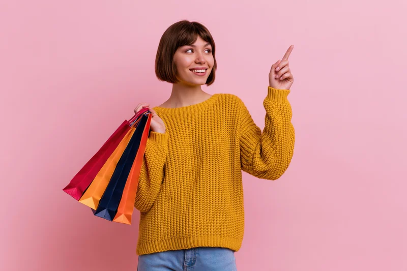 Young woman holding colorful shopping bags and pointing upward against pink background