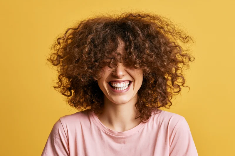 Woman laughing with voluminous curly brown hair covering her face against yellow background