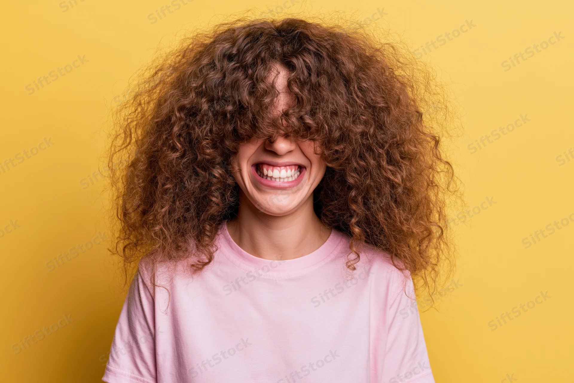 Young woman with voluminous curly brown hair laughing joyfully in front of yellow background