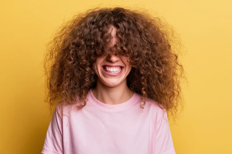 Young woman with voluminous curly brown hair laughing joyfully in front of yellow background