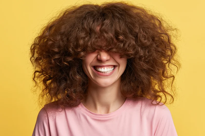 Woman with voluminous curly brown hair smiling against yellow background wearing pink shirt