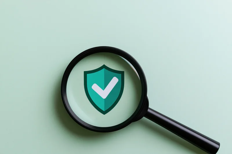 Black magnifying glass over mint-green surface showing shield with checkmark symbol
