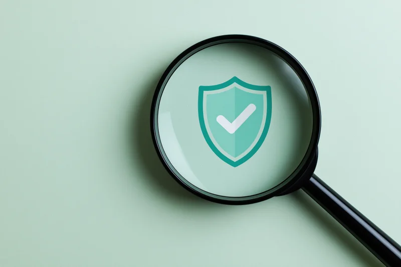 Black magnifying glass on mint-green surface showing shield with checkmark symbol