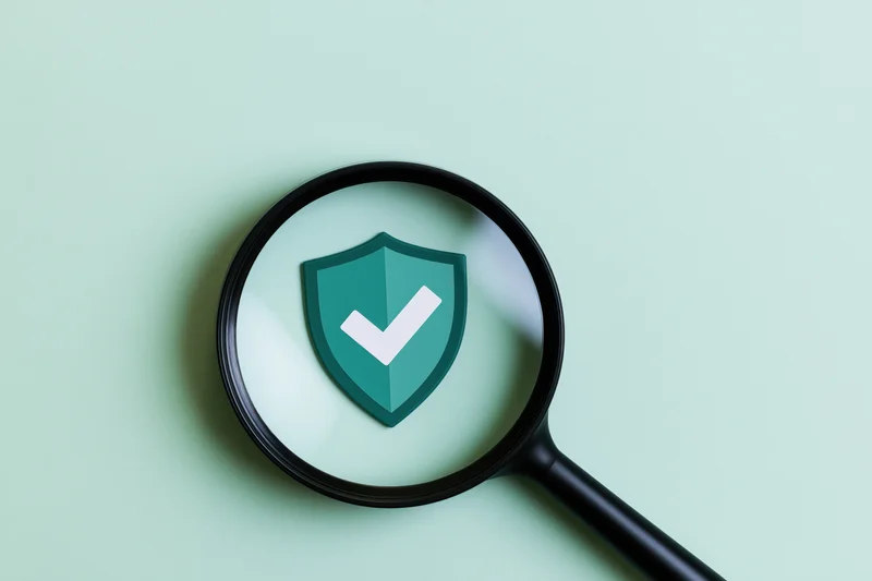 Magnifying glass over a green shield with white checkmark on mint surface