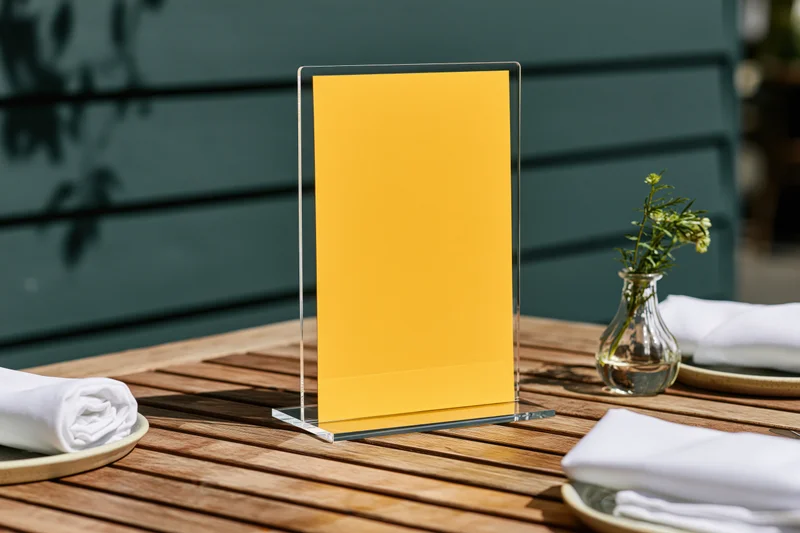 Bright yellow menu card in clear acrylic stand on wooden table with plates and napkins outdoors