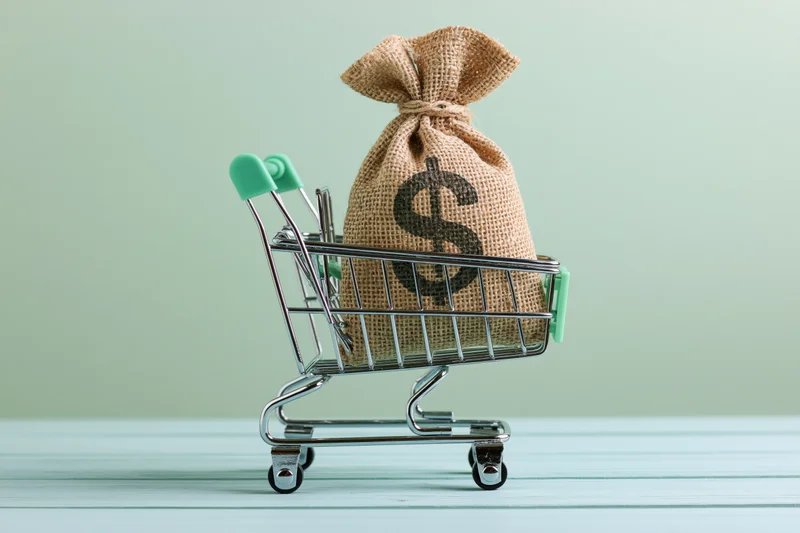 Burlap money bag with dollar sign in miniature shopping cart on wooden surface