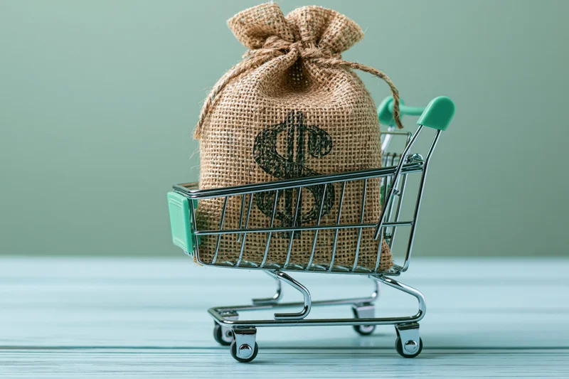 Burlap money bag with dollar sign in miniature shopping cart on wooden surface