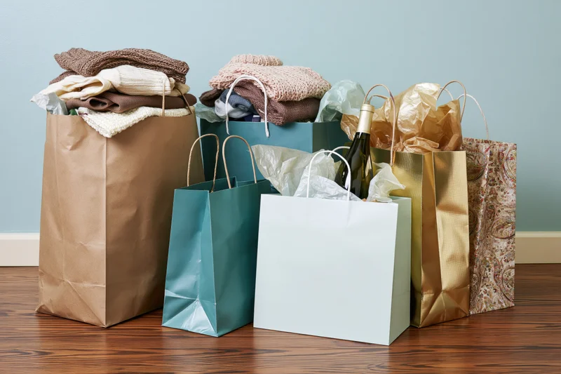 Assorted shopping bags filled with clothing and a wine bottle arranged on a wooden floor