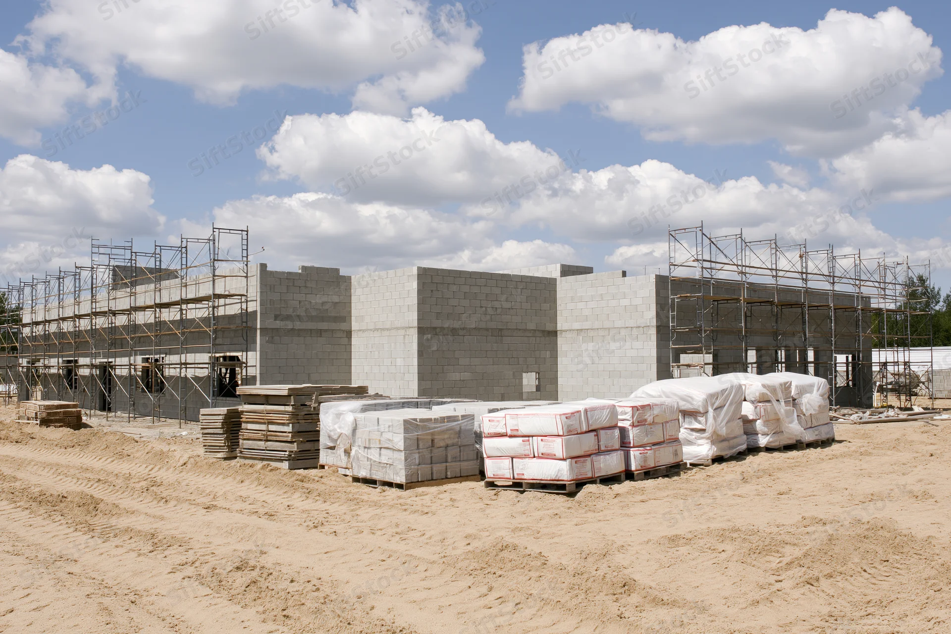 Concrete block building under construction with scaffolding and stacked materials on a sandy site