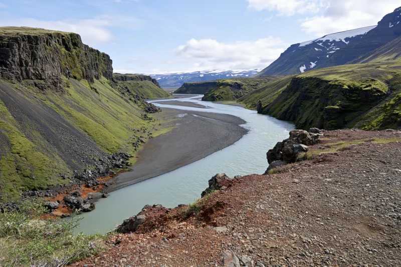 Glacial river winding through steep icelandic highland valley with mossy cliffs and snow-capped mountains in background