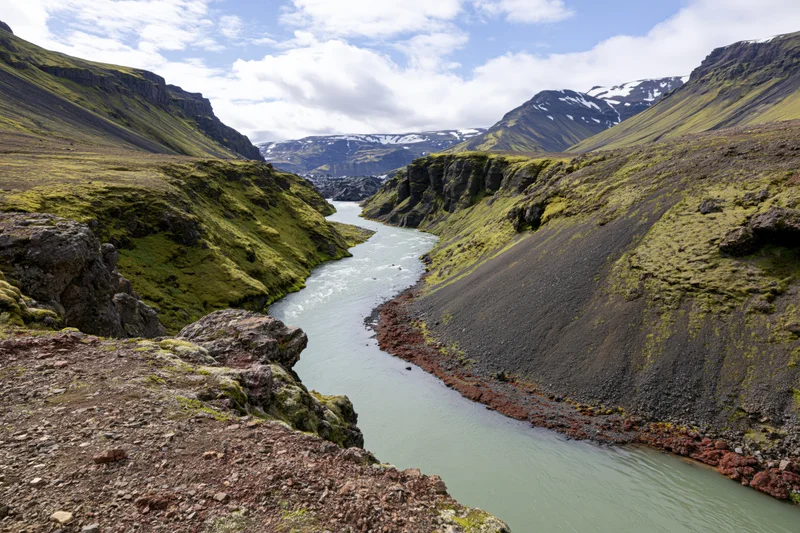 Glacial river winding through steep icelandic highland valley with moss-covered slopes and snow-capped mountains in background