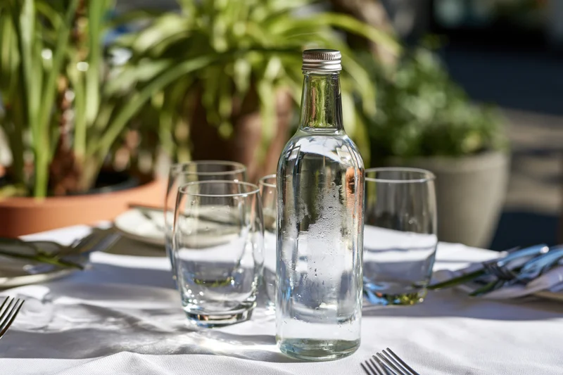 Glass water bottle with metal cap on set outdoor dining table with glassware and potted plants