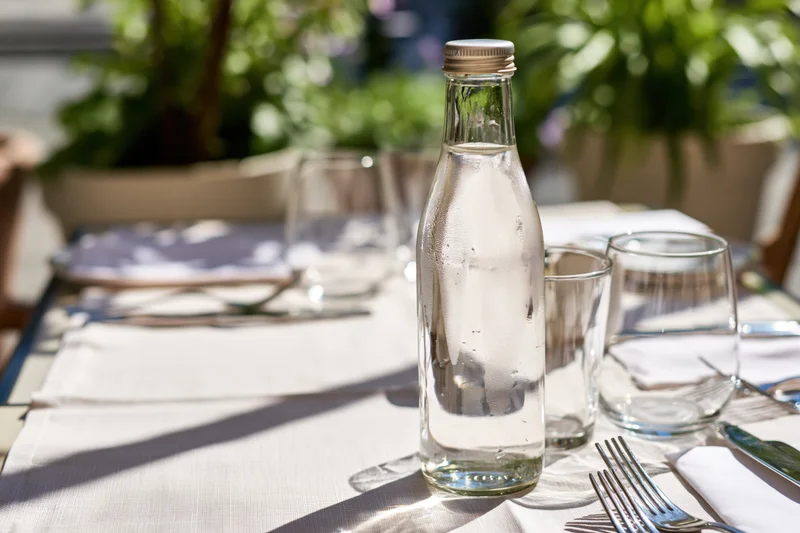 Glass water bottle with metal cap on set outdoor restaurant table with cutlery and napkins under sunlight