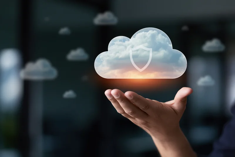Hand holding a glowing translucent cloud with a shield symbol over a dark office background