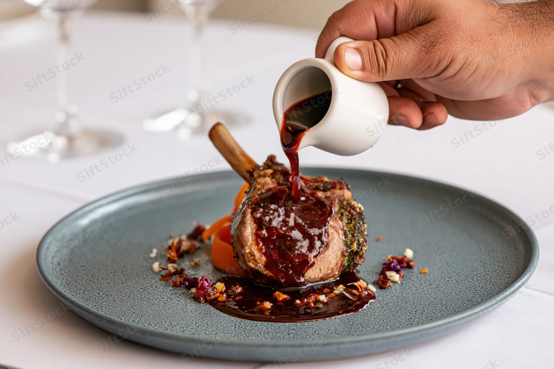Hand pouring dark red jus from a small white ceramic pitcher onto a lamb chop on a gray plate at a fine dining table