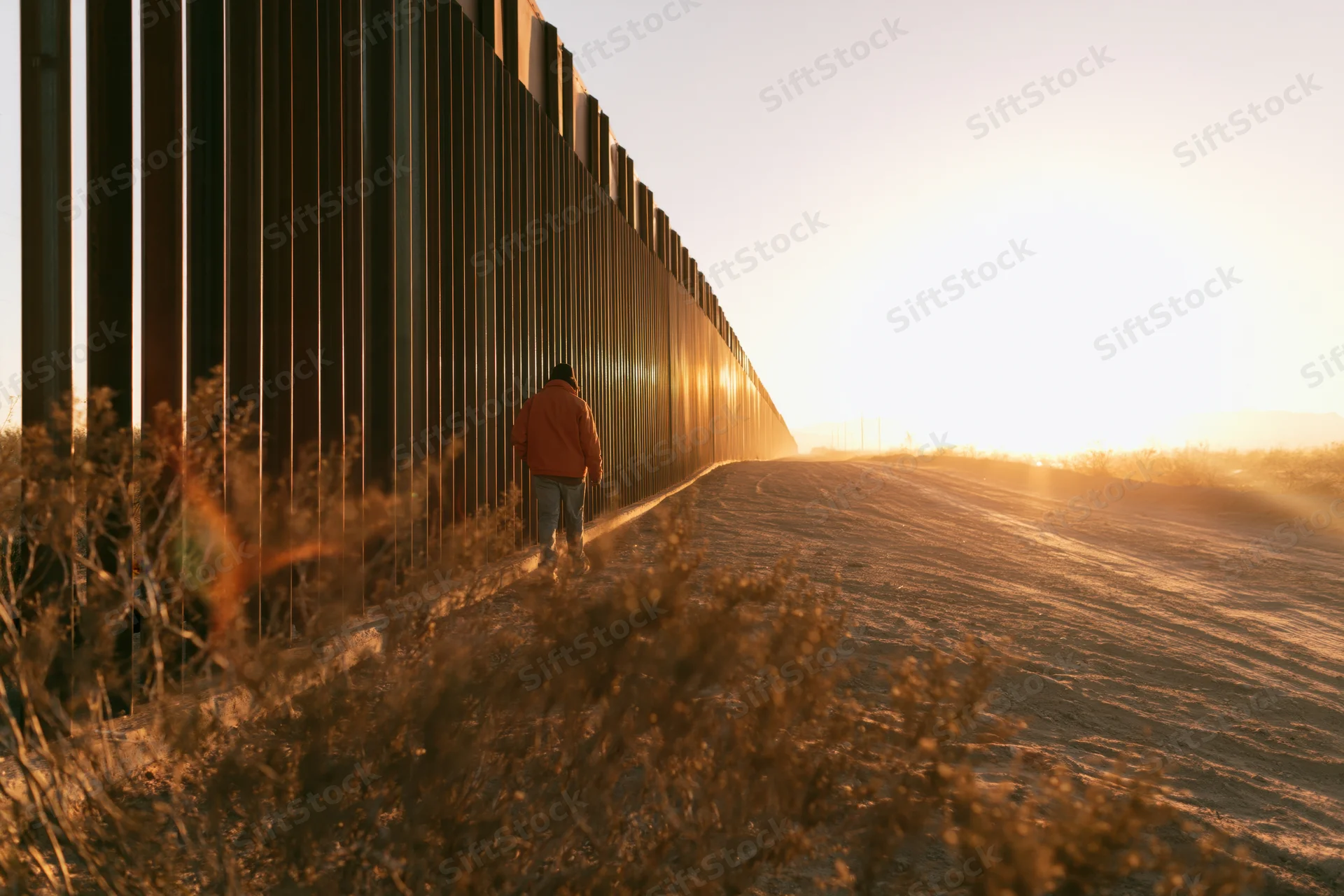 Man walking along a metal border fence in a desert at sunset