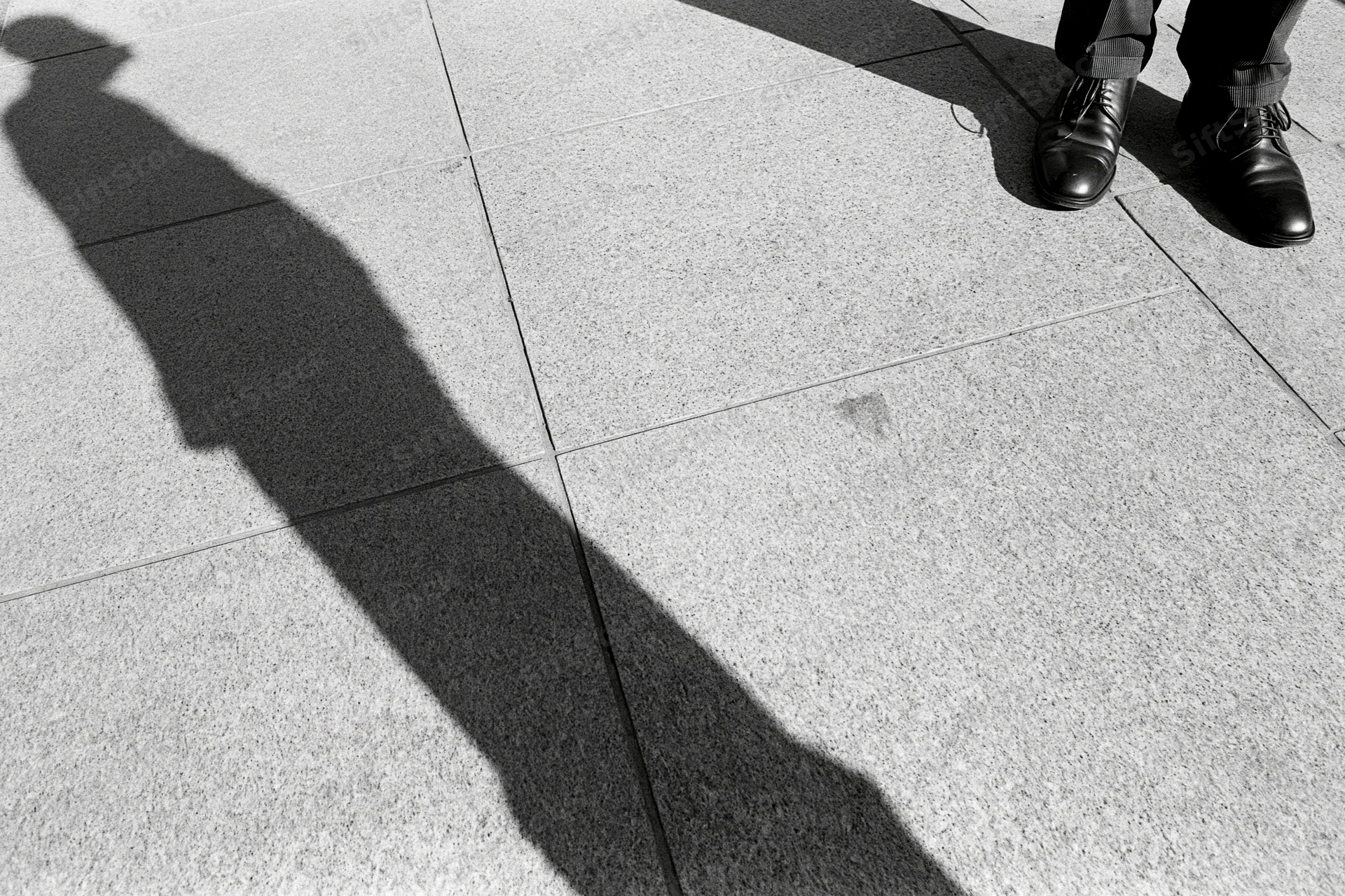 Long shadow of a standing man cast across grey pavement in urban setting
