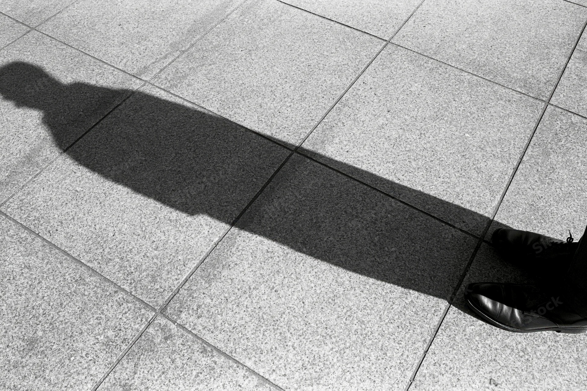 Long shadow of a standing person cast across tiled pavement in black and white